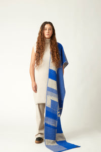 Wide shot of a young woman with a knit scarf draped over the shoulder. She's wearing a neutral toned outfit and black shoes. Packhshot of a jacquard knit scarf in cobalt blue and beige. The scarf has an abstract geometric bitmap pattern.
